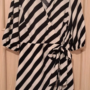 Svartvit randig omlottklänning från F&F - Snygg svartvit randig omlottklänning från F&F med knytband i midjan. Klänningen har trekvartsärmar och ett stilrent mönster som passar perfekt för olika tillfällen.