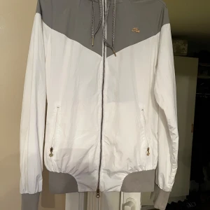 Vit och grå vindjacka från Nike - Snygg vit och grå vindjacka från Nike med dragkedja och justerbar huva. Jackan har två fickor med dragkedjor och ett litet Nike Air-märke på bröstet. Perfekt för en sportig look.