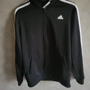Svart träningsjacka från Adidas - Snygg svart träningsjacka från Adidas med vita ränder längs ärmarna och dragkedja framtill. Perfekt för sportiga aktiviteter eller en avslappnad stil. Adidas-logga på bröstet ger en klassisk touch.