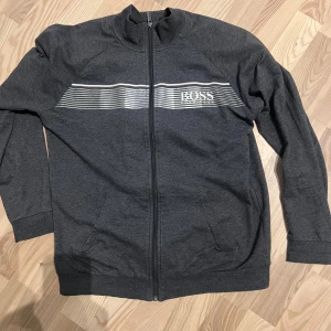 Mörkgrå tröja med dragkedja från BOSS - Säljer en stilren mörkgrå tröja från BOSS med dragkedja och ett vitt randigt mönster över bröstet. Tröjan har långa ärmar och en hög krage, perfekt för en avslappnad look.