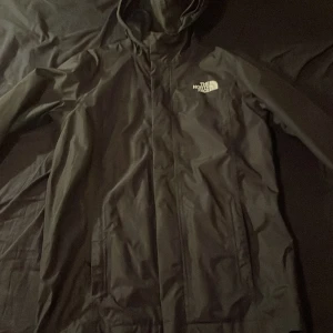 Svart regnjacka från The North Face - Säljer en svart regnjacka från The North Face med huva och dragkedja. Perfekt för regniga dagar och har en stilren design med logotyp på bröstet. Möts endast!