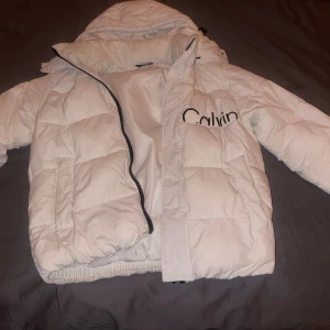 Vit pufferjacka från Calvin Klein - Säljer en snygg vit pufferjacka från Calvin Klein. Jackan har en dragkedja framtill och en stor logga på bröstet och ryggen. Perfekt för kyliga dagar med sin vadderade design och huva. Ordinarie pris 2750kr