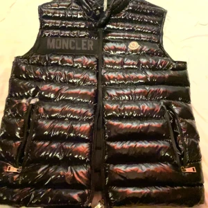 Svart dunväst från Moncler  - Snygg svart dunväst från Moncler med glansig finish och dragkedja framtill. Västen har två fickor med dragkedjor och Moncler-logga på bröstet. Perfekt för kyligare dagar.