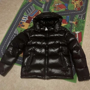 Svart pufferjacka från Moncler - Snygg svart pufferjacka från Moncler med glansig finish och dragkedja. Jackan har en huva och långa ärmar, perfekt för kyliga dagar. Den quiltade designen ger extra värme och stil.