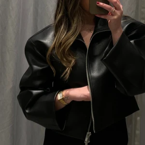 Populär Svart skinnjacka - Snygg svart skinnjacka med dragkedja och croppad design. Jackan har långa ärmar och en stilren look, perfekt för en trendig outfit. Halskrage och grövre material och dragkedja 