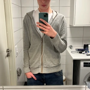 Grå hoodie med dragkedja - Säljer en stilren grå Massimo dutti hoodie med dragkedja framtill. Perfekt för en avslappnad look. Den har långa ärmar och en bekväm huva. Storlek L men sitter som M modellen är 190 och bär vanligtvis M