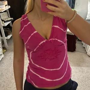 Rosa ärmlös topp med batikmönster - Säljer en snygg rosa ärmlös topp med vitt batikmönster och v-ringning. Toppen har en dekorativ broderad detalj framtill. Perfekt för sommardagar!