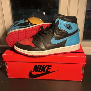 Nike Air Jordans 1 - Säljer ett par Nike Air Jordans i en cool färgkombination av svart, blå och röd. Skorna har en klassisk hög design med snörning och en vit sula. Perfekta för sneaker-entusiaster!