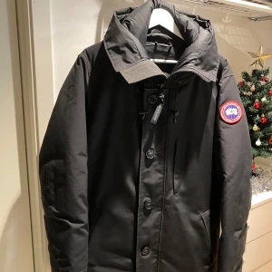 Svart dunjacka från Canada Goose - Säljer en svart dunjacka från Canada Goose med huva och dragkedja. Jackan har knappar framtill och en broderad logotyp på ärmen. Har fickor inuti jackan där med.  Använd en halv vinter ungefär och är forfarande fräsch som ny. Perfekt för kalla vinterdagar.