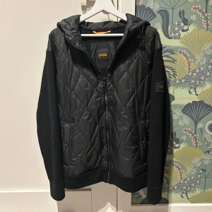 Svart cardigan från Boss - Snygg svart quiltad jacka från Boss med huva och dragkedja. Jackan har ribbade ärmar och en stilren design, perfekt för kyligare dagar. Den har en diskret logga på ärmen och en bekväm passform. Använd ett fåtal gånger men ser ut som ny.