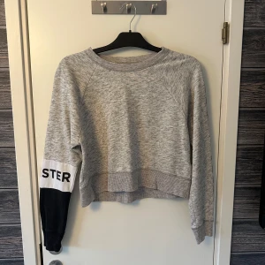 Grå sweatshirt från Hollister - Säljer en grå sweatshirt från Hollister med textdetaljer på ärmarna. Tröjan har en avslappnad passform och är perfekt för en casual look. Den har långa ärmar och ribbade muddar vid ärmslut och nederkant.