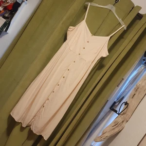 Linneklänning -  Benvit/beige linneklänning med knappar framtill och smala axelband. Klänningen har en smockdetalj på ryggen för extra komfort och passform. Perfekt för varma dagar. Knapparna går ej att öppna/stänga/knäppa upp. Uppskattar den till en storlek LARGE. 