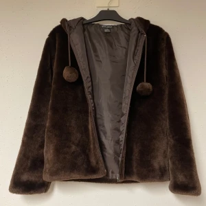 Brun pälsjacka från Caramelle - Mysig brun pälsjacka från Caramelle med huva och dekorativa tofsar. Jackan har en mjuk yta och är perfekt för kyligare dagar. Den är tillverkad i akryl och polyester med ett foder av polyester. (Passar för folk som är mindre också då den är liten i storleken. Jag skulle säga att en S eller M skulle också kunna ha den🫶🏻💗)