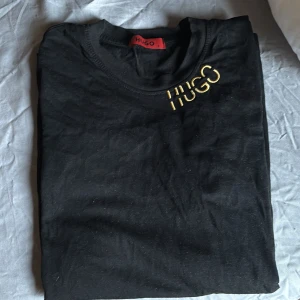 Svart t-shirt från Hugo - Snygg svart t-shirt från Hugo med guldfärgad logga på bröstet. Perfekt för en stilren look. Kortärmad och tillverkad i mjukt material för extra komfort.
