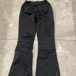 Svarta skidbyxor med utsvängda ben - Snygga svarta byxor med bootcut-stil och hög midja. Byxorna har justerbar midja och dekorativa sömmar över knäna. Perfekta för en trendig look i skidbacken.