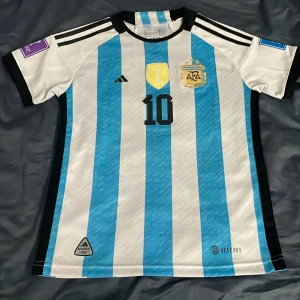 Argentina fotbollströja med nummer 10 - Säljer en officiell Argentina fotbollströja från Adidas med nummer 10 och namn Messi på ryggen. Tröjan har de klassiska blå och vita ränderna och AFA-emblemet på bröstet. Kortärmad design med autentiska detaljer från 2022.       Kom gärna med förslag 