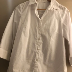 Vit skjorta med trekvartsärm - Klassisk vit skjorta med trekvartsärm och knappar framtill. Perfekt för en stilren look. Skjortan har en enkel design med krage och är tillverkad i ett lätt och bekvämt material.