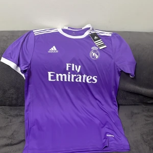 Lila Real Madrid fotbollströja - Snygg lila Real Madrid tröja med 'Fly Emirates' tryck på framsidan och 'Ronaldo 7' på baksidan. Tröjan har korta ärmar och vita detaljer vid krage och ärmslut. Perfekt för fotbollsfans!