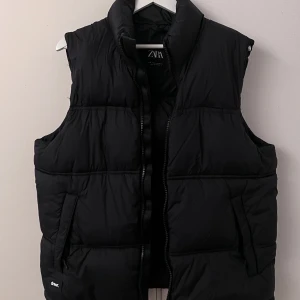Svart dunväst från Zara - Snygg svart dunväst från Zara med dragkedja framtill. Perfekt för kyliga dagar och enkel att matcha med olika outfits. Västen har en stilren design med hög krage och praktiska fickor.