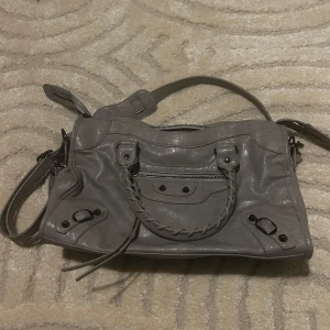 Grå handväska med flätad detalj - Snygg grå handväska flätad detalj på framsidan och nitar. Väskan har även ett långt band som går att ta av och på och flera praktiska fack inuti. Den är runt 30x25cm så en perfekt skolväska som en dator får plats i!💋har andvänt en gång så den är i nyskick