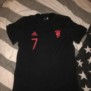 Svart fotbollströja från Adidas - Snygg svart fotbollströja från Adidas med röd nummer 7 och klubbemblem på framsidan. Namnet 'Ronaldo' och nummer 7 är tryckt på baksidan. Perfekt för fotbollsfans!