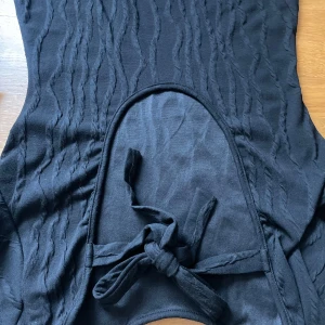 BUDA: Lång svart elegant klänning med öppen rygg - Denna klänning sitter som en smack på kroppen, den är lång och har eleganta mönster längst klänningen. Den öppna ryggen har band, man väljer själv hur tajt den ska sitta.