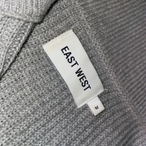 Grå stickad tröja från East West - Säljer en stilren grå stickad tröja från East West. Tröjan har en dragkedja framtill och en hög krage som ger extra värme. Perfekt för kyliga dagar! Säljer den eftersom den inte passar mig är 176
