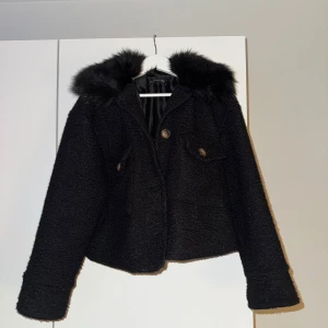 Svart teddyjacka med pälskrage - Snygg svart teddyjacka med pälskrage och knappar framtill. Perfekt för kyliga dagar med sin mysiga och varma design. Jackan har långa ärmar och en croppad passform.