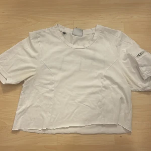 Vit croppad t-shirt från Gymshark - Snygg vit croppad t-shirt från Gymshark med coolt tryck på baksidan i svart och grönt. T-shirten har korta ärmar med en praktisk ficka med dragkedja. Perfekt för en avslappnad stil.