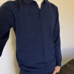 Mörkblå pikétröja  - Snygg mörkblå pikétröja med krage och knappar framtill. Perfekt för en stilren look. Tröjan har långa ärmar och en liten metallbricka med logotyp vid nederkanten. Den är i storlek M och modellen är 180 cm lång. Det är bara att höra av sig vid frågor eller funderingar