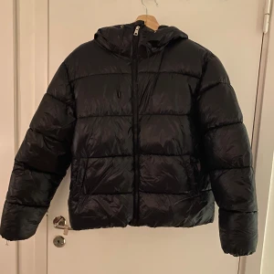 Svart pufferjacka med huva - Säljer en svart pufferjacka med huva. Jackan har en dragkedja framtill och är perfekt för kyligare dagar. Den är långärmad och har en klassisk pufferdesign som ger både stil och värme.