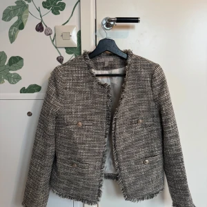 Grå kavaj från H&M - Snygg grå kavaj från H&M i ett tweed-liknande material. Kavajen har en fransig kant och knappar framtill. Perfekt för en stilren look.