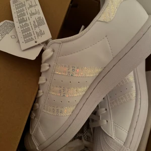 Vita Adidas Superstar sneakers - Snygga vita Adidas Superstar sneakers med glittriga detaljer på de klassiska tre ränderna. Skorna har snörning och en ikonisk tåhätta. Perfekta för en stilren look.