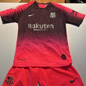 FC Barcelona fotbollströja med shorts - Snygg FC Barcelona fotbollströja i svart och knallorange med korta ärmar och matchande shorts. Tröjan har tryck med Rakuten och Messi 10 på ryggen. Perfekt för fotbollsfans!