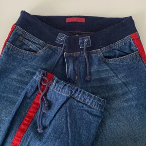 Blå jeans med röd rand - Snygga blå jeans med en iögonfallande röd rand längs sidorna. Byxorna har en bekväm resårmidja med snörning och klassiska femficksdetaljer. Perfekta för en avslappnad stil med en twist.
