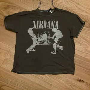 Grå Nirvana t-shirt - Cool grå t-shirt med Nirvana-tryck i vitt. T-shirten har ett rockigt motiv med bandmedlemmar i aktion. Perfekt för fans av klassisk grunge. Kortärmad och i normal passform. Tröjan har lite fläckar som knappt är synliga för ögat.