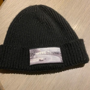 Svart mössa med ett motiv - Snygg svart ribbad mössa med en unik broderad patch framtill. Perfekt för att hålla värmen under kyliga dagar och ge en stilren touch till din outfit.