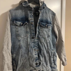 Jeansjacka med grå ärmar och huva - Snygg jeansjacka från Young & Reckless med grå ärmar och huva. Jackan har knappar framtill och två bröstfickor med lock. Perfekt för en avslappnad stil. Behöver strykas bara