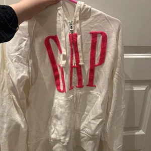 Vit hoodie från GAP - Säljer en vit hoodie från GAP med dragkedja och stor rosa logga på framsidan. Perfekt för en avslappnad stil. Den har en klassisk passform och är tillverkad i mjukt material. Aldrig använd.