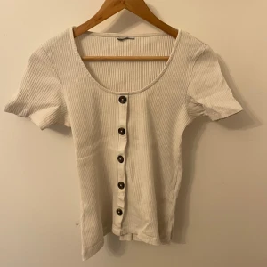 Vit ribbad topp med knappar - Säljer en stilren vit ribbad topp med korta ärmar och knappar framtill. Perfekt för en avslappnad look.