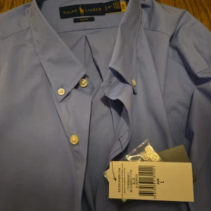 Blå skjorta från Ralph Lauren - Snygg blå skjorta från Ralph Lauren med klassisk krage och knappar framtill. Perfekt för en stilren look. Skjortan är i ett mjukt material och har en tidlös design.