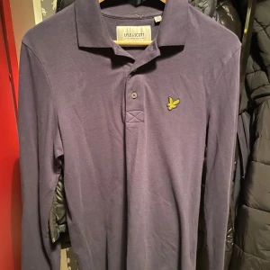 Mörkblå pikétröja från Lyle & Scott - Snygg mörkblå pikétröja från Lyle & Scott med klassisk krage och knappslå. Tröjan är ny och är endast andvänd 3-4 gånger, Tröjan har ett broderat emblem på bröstet och är tillverkad i ekologiskt material. Perfekt för en stilren look. Nypris 749, Hör av dig vid Funderingar🙌