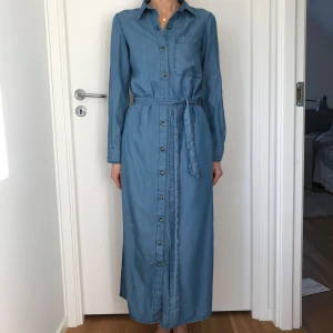 Zara denim klänning Stl XS Nyskick  - Lång blå denimklänning med knappar framtill och bälte i midjan. Klänningen har slits i sidorna. 100% Tencel för mjukt och fint fall, mkt skön kvalitet att ha på sig. Storlek XS men passar en normal small bra. Bara använd en gång, är i nyskick. Nypris 699:- Rök o djurfritt hem.
