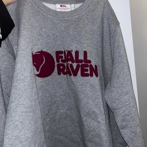 Grå sweatshirt från Fjällräven - Säljer en grå sweatshirt från Fjällräven med deras ikoniska logga i vinrött på bröstet. Tröjan har en klassisk rund halsringning och är perfekt för en avslappnad stil.
