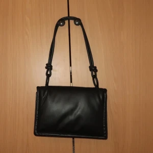 Svart axelväska i skinn - Snygg svart axelväska i skinn med enkel design. Väskan har ett lock med magnetknappar och flera fack inuti för smidig organisering. Perfekt för att bära dina dagliga nödvändigheter med stil.