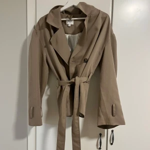 Beige trenchcoat från Saint - Snygg beige trenchcoat från Saint med knytbälte i midjan och dubbelknäppning. Jackan har långa ärmar och en klassisk krage, perfekt för höst och vår. En stilren och tidlös design som passar till många olika outfits.