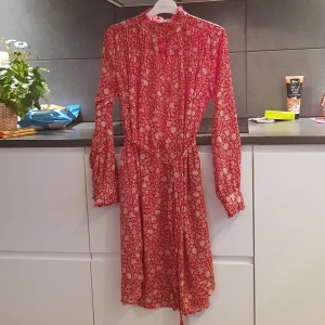 Röd blommig klänning från Holly & Whyte - Charmig röd klänning med blommigt mönster från Holly & Whyte by Lindex. Klänningen har långa ärmar och en knytning i midjan för en smickrande passform. Perfekt för en somrig look!