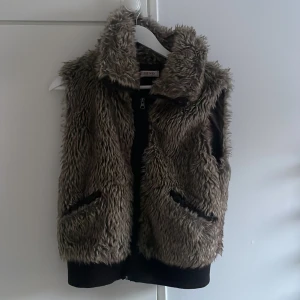 Brun pälsväst från Orsay - Säljer en snygg brun pälsväst från Orsay med dragkedja framtill. Västen har en mysig och fluffig look, perfekt för kyligare dagar. Den är ärmlös och har en hög krage för extra värme. Vet inte storlek men passar xs/s/m