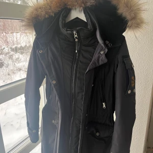 Svart parkajacka med pälsdetaljer från Wiggy's - Snygg och funktionell svart parkajacka från Wiggy's med laminerad isolering. Den har som en inbyggd väst/jacka. Jackan har en avtagbar huva med äkta pälsdetaljer och flera praktiska fickor. Perfekt för kalla vinterdagar. Den är märkt med Stl 1. Jag skulle säga XS-S 