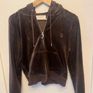 Brun velour hoodie från Juicy Couture - Säljer en brun velour hoodie från Juicy Couture med dragkedja och huva. Tröjan har långa ärmar och en klassisk känguruficka framtill. Perfekt för en avslappnad och stilren look.